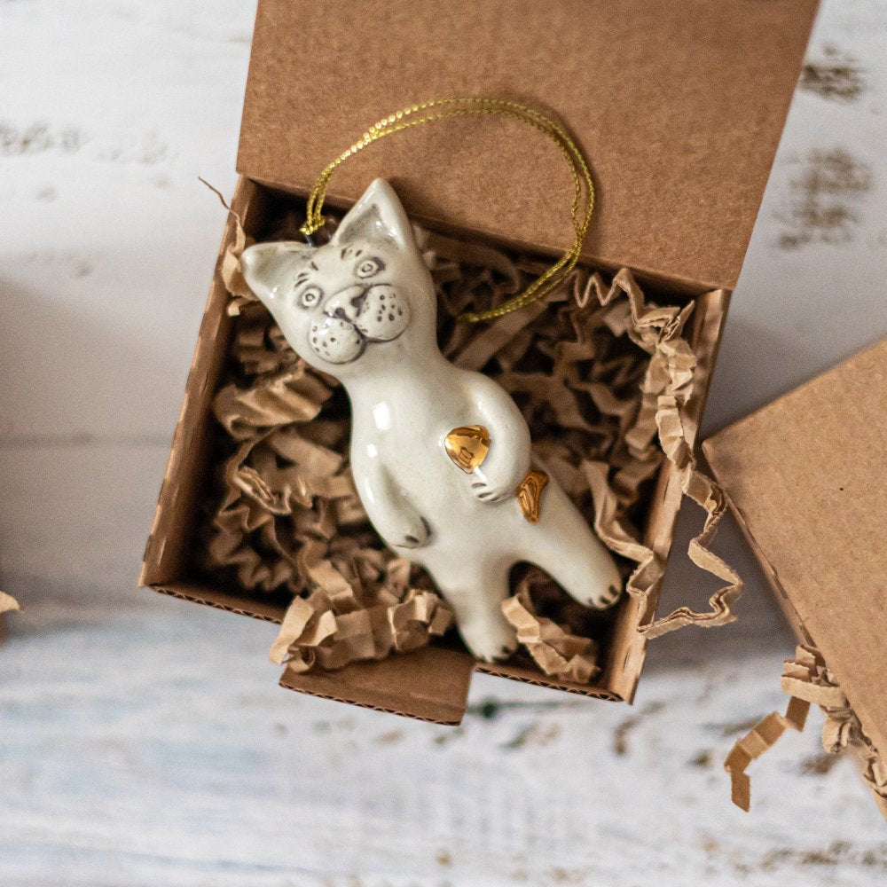 Ceramic cat ornament with hand painted mouth, nose and ears holding a golden fish. The decoration is white with black markings and is hanging from a gold string.