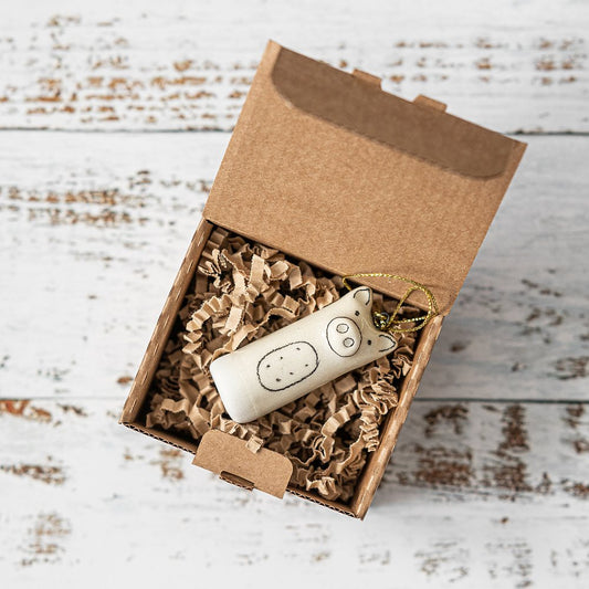 A white ceramic pig ornament with black painted eyes and a nose. The decoration is white with black markings and is hanging from a gold string.