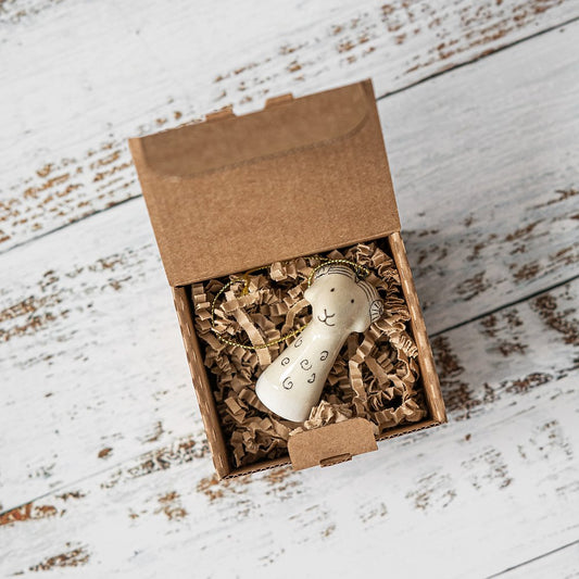 A white ceramic sheep ornament with black painted eyes and a small mouth. The decoration is white with black markings and is hanging from a gold string.