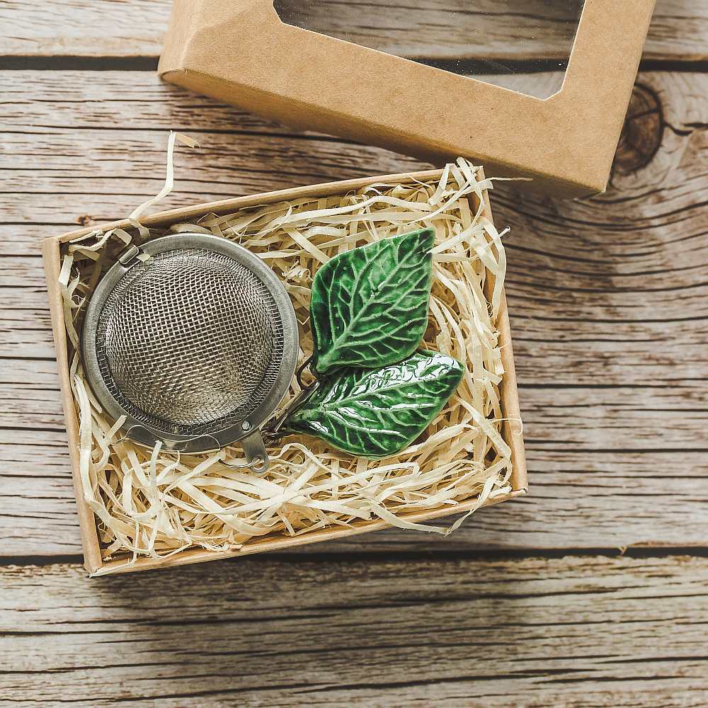 Tea infuser with ceramic green leaves hanging from the chain. Packed in a paper gift box.