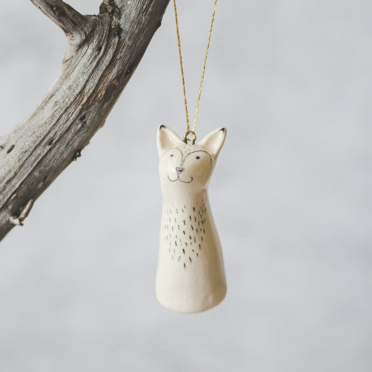 A smiling ceramic fox ornament with hand-drawn black eyes, mouth and a belly. The decoration is white with black markings and is hanging from a gold string.