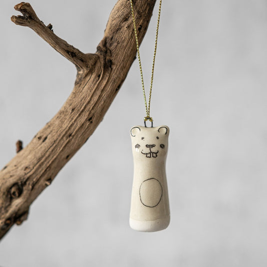 A white ceramic beaver ornament with black painted eyes and a smiling mouth. The decoration is white with black markings and is hanging from a gold string.