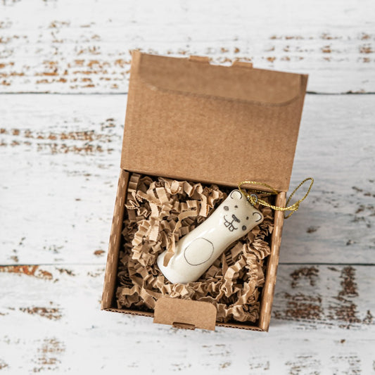 A white ceramic beaver ornament with black painted eyes and a smiling mouth. The decoration is white with black markings and is hanging from a gold string.