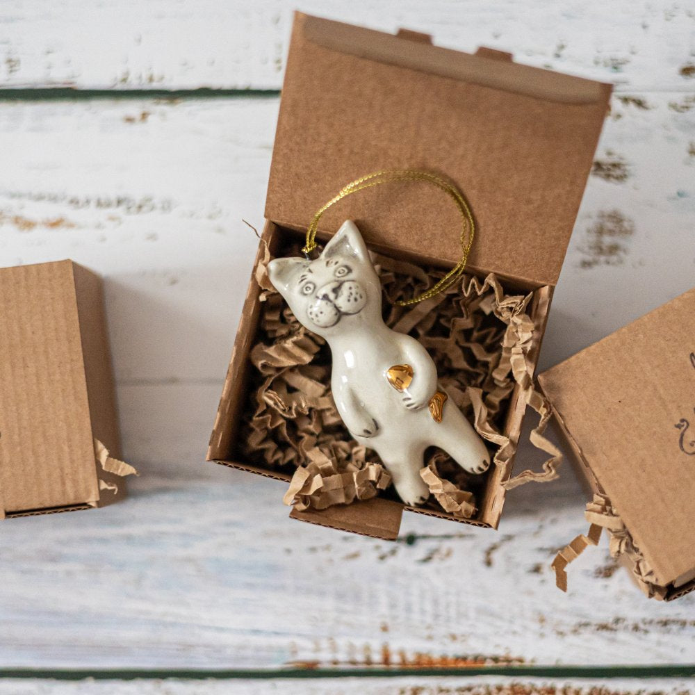 Ceramic cat ornament with hand painted mouth, nose and ears holding a golden fish. The decoration is white with black markings and is hanging from a gold string.