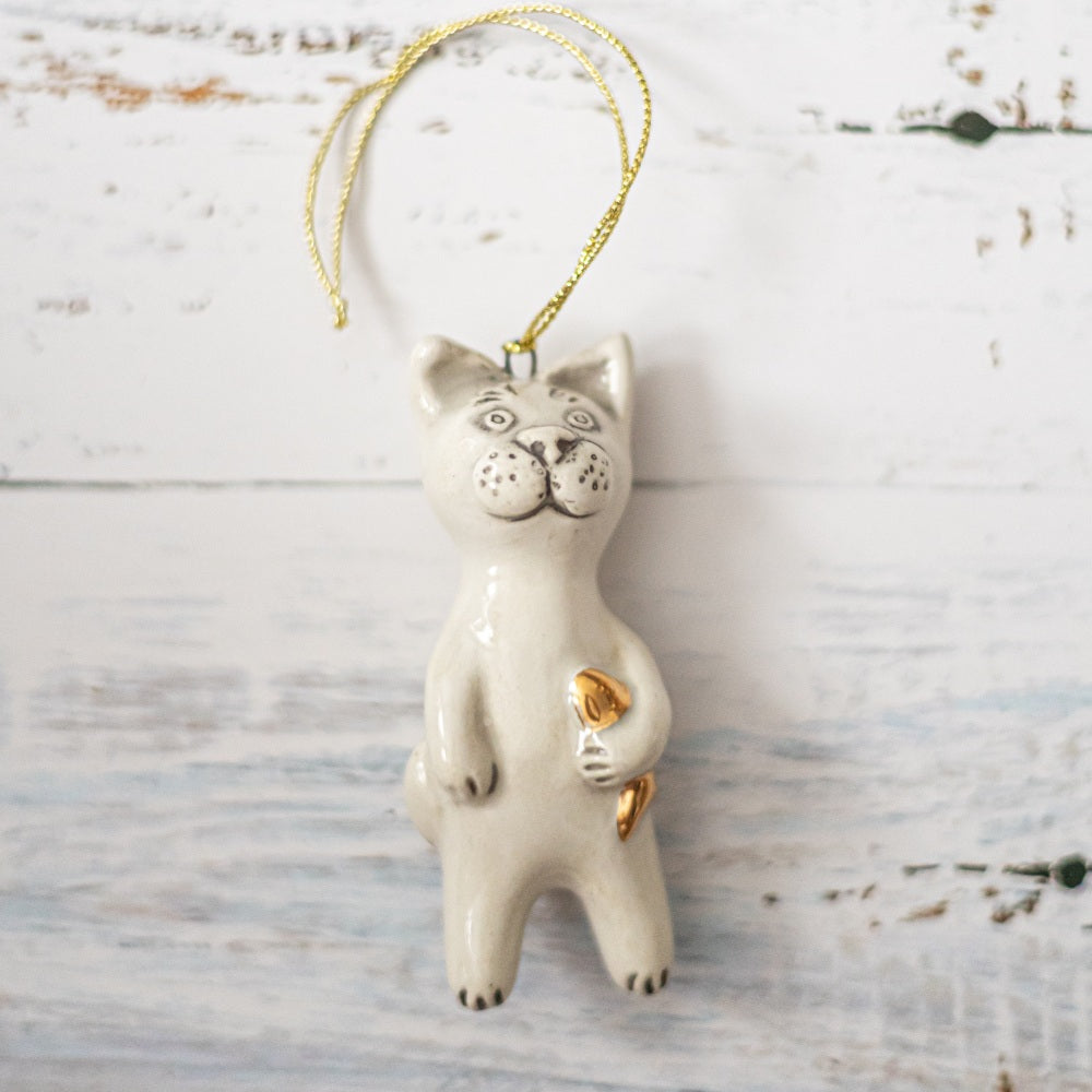 Ceramic cat ornament with hand painted mouth, nose and ears holding a golden fish. The decoration is white with black markings and is hanging from a gold string.