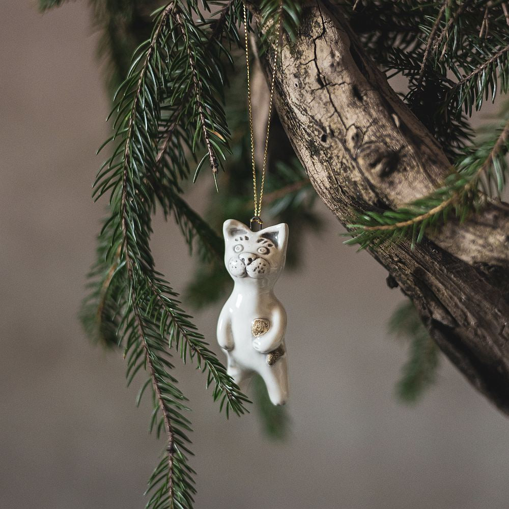 Ceramic cat ornament with hand painted mouth, nose and ears holding a golden fish. The decoration is white with black markings and is hanging from a gold string.