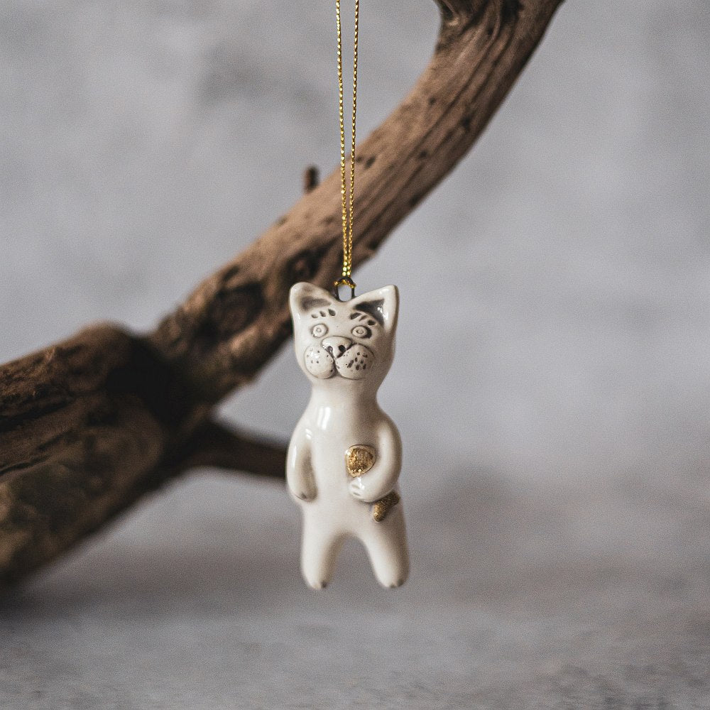 Ceramic cat ornament with hand painted mouth, nose and ears holding a golden fish. The decoration is white with black markings and is hanging from a gold string.