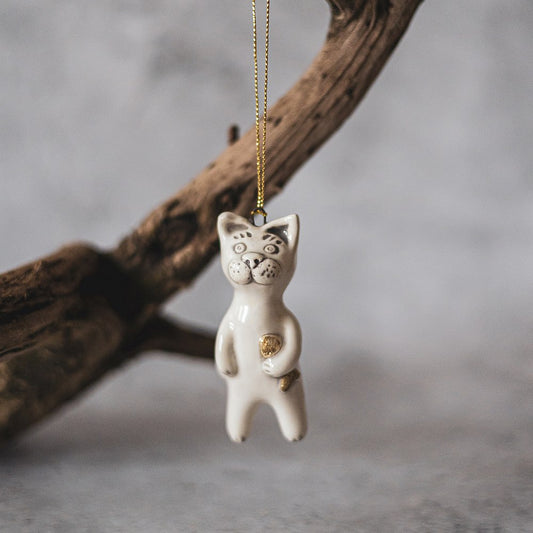 Ceramic cat ornament with hand painted mouth, nose and ears holding a golden fish. The decoration is white with black markings and is hanging from a gold string.