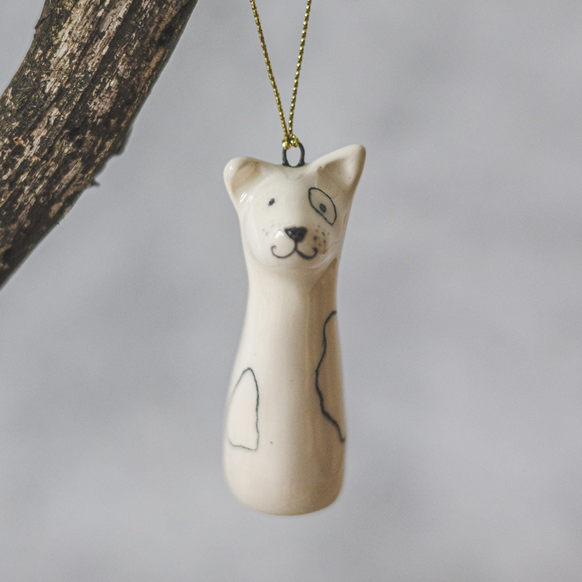 A smiling ceramic dog ornament with hand-drawn black eyes, mouth and a black nose. The decoration is white with black markings and is hanging from a gold string.