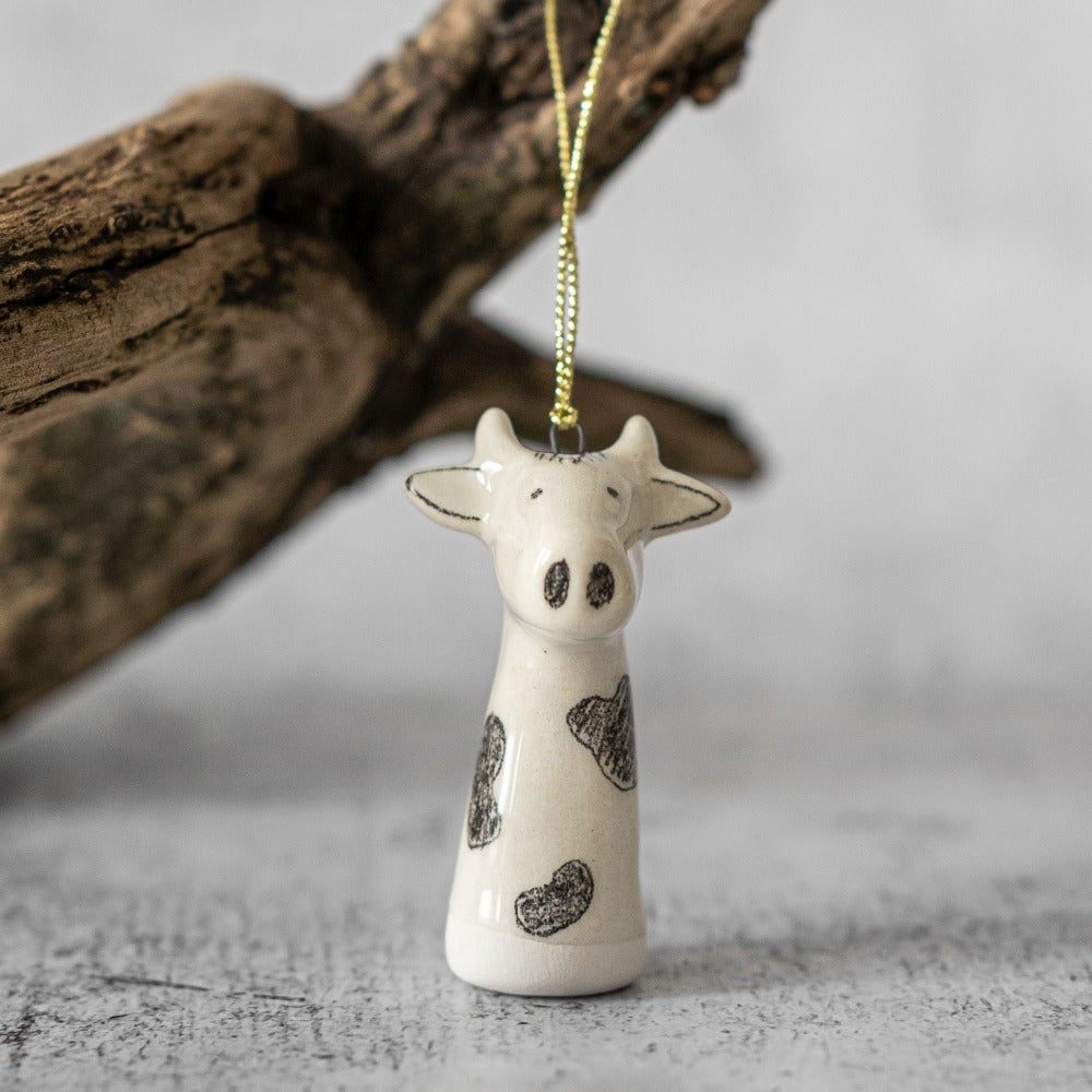 A ceramic cow ornament with black painted eyes and nose. The decoration is white with black spots and is hanging from a gold string.