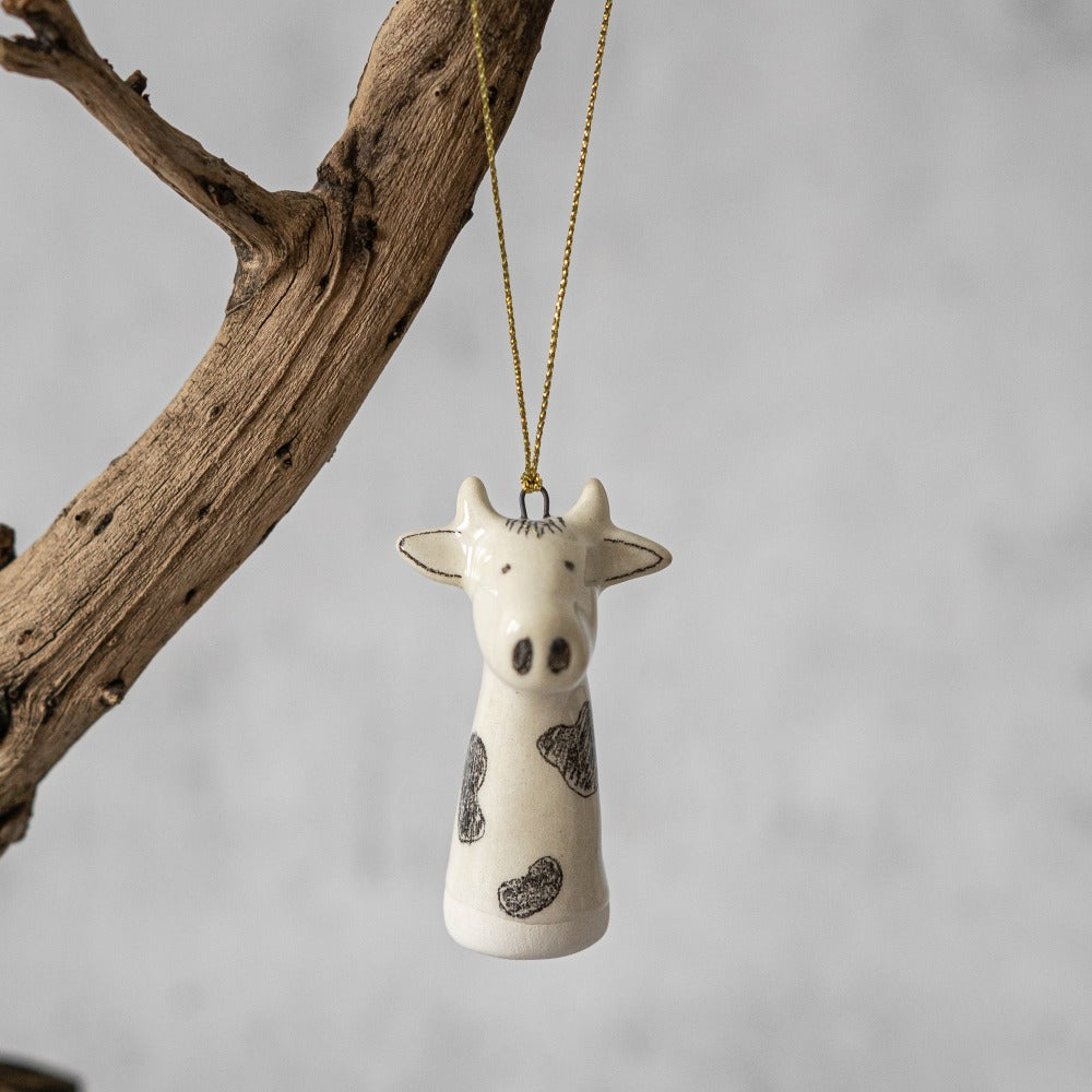 A ceramic cow ornament with black painted eyes and nose. The decoration is white with black spots and is hanging from a gold string.