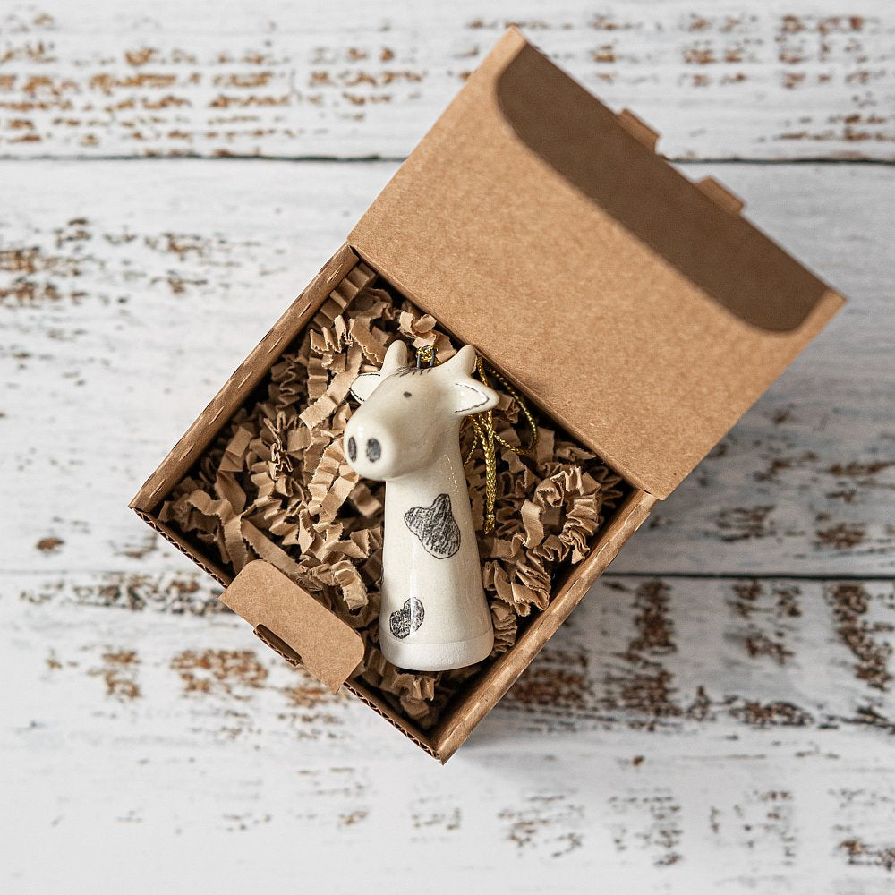 A ceramic cow ornament with black painted eyes and nose. The decoration is white with black spots and is hanging from a gold string.