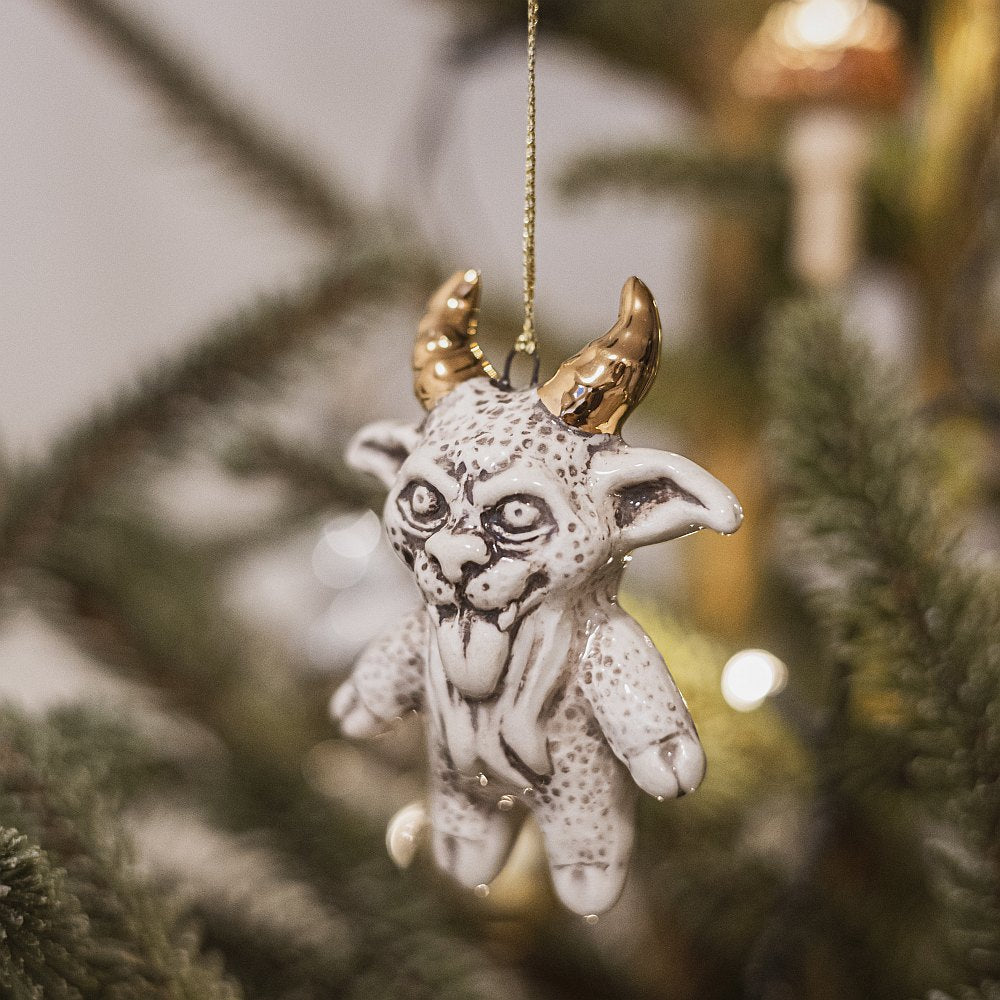 Ceramic devil ornament with golden horns. The decoration is white with black markings and is hanging from a gold string.