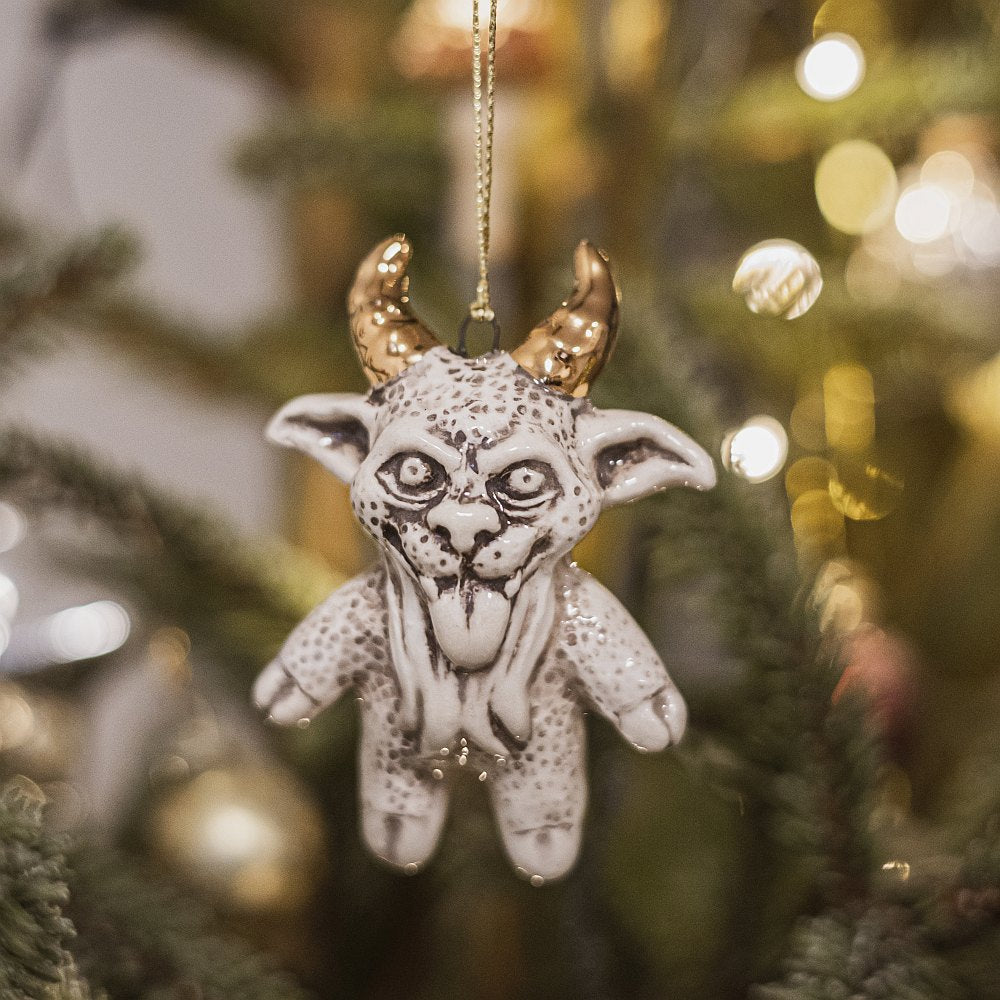 Ceramic devil ornament with golden horns. The decoration is white with black markings and is hanging from a gold string.