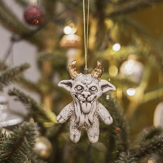 Ceramic devil ornament with golden horns. The decoration is white with black markings and is hanging from a gold string.