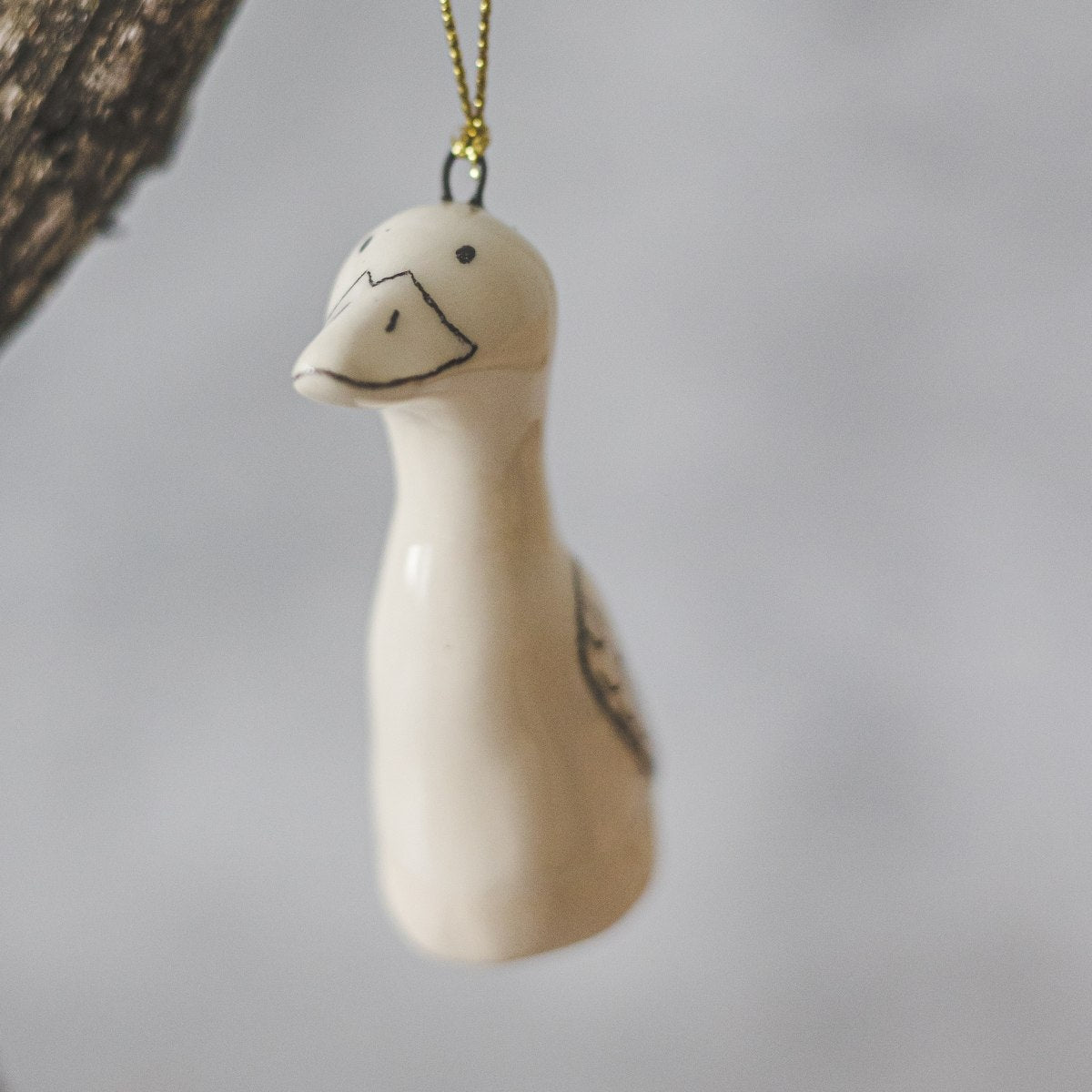 A ceramic duck ornament with hand-drawn black eyes, beak and wings. The decoration is white with black markings and is hanging from a gold string.