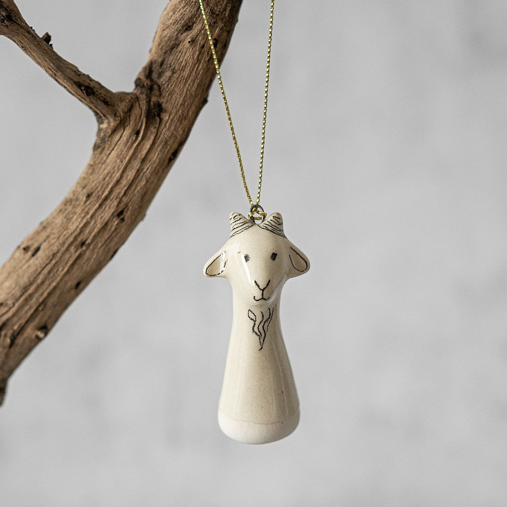 A white ceramic goat ornament with black painted eyes and a small mouth. The decoration is white with black markings and is hanging from a gold string.