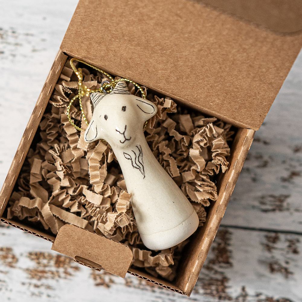 A white ceramic goat ornament with black painted eyes and a small mouth. The decoration is white with black markings and is hanging from a gold string.