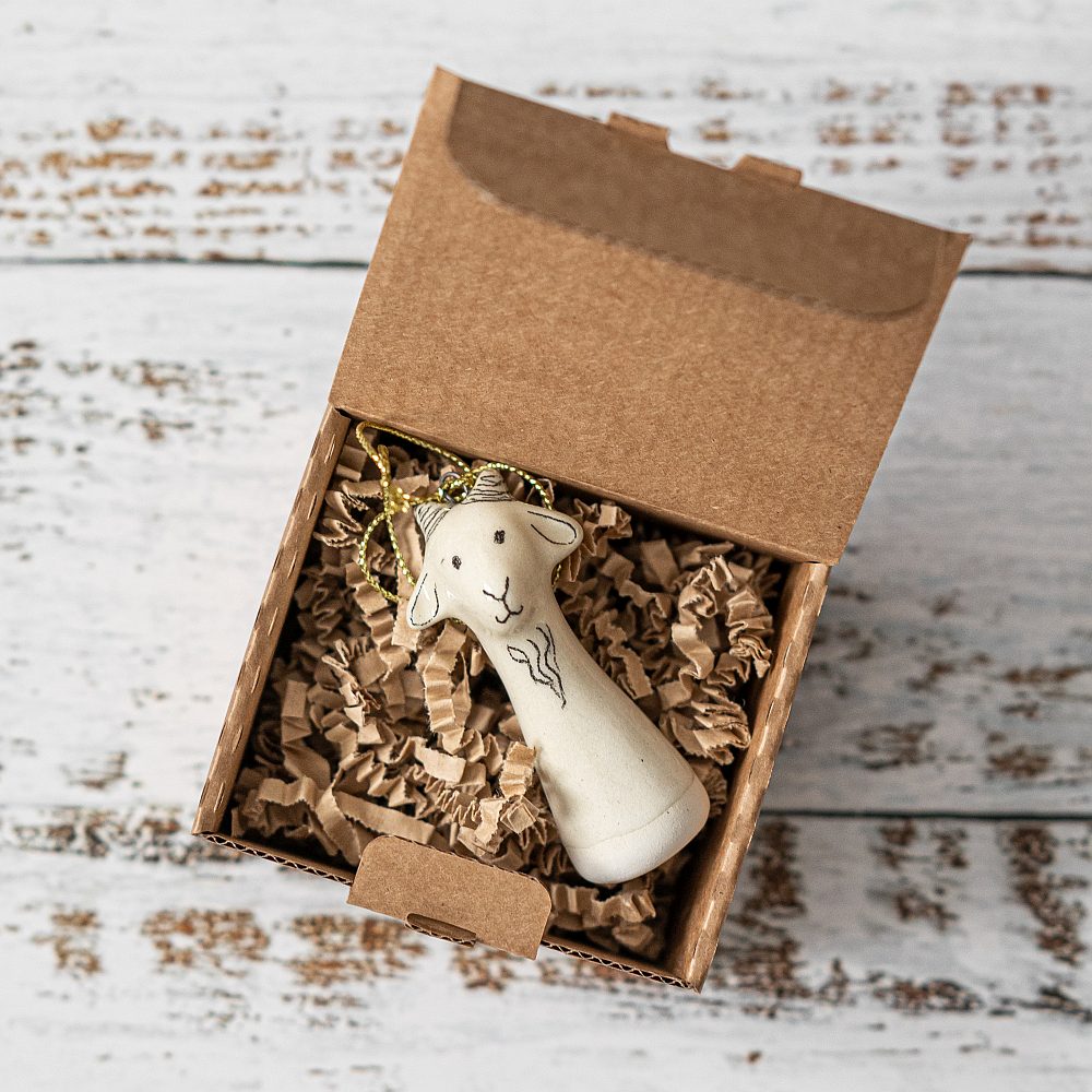 A white ceramic goat ornament with black painted eyes and a small mouth. The decoration is white with black markings and is hanging from a gold string.