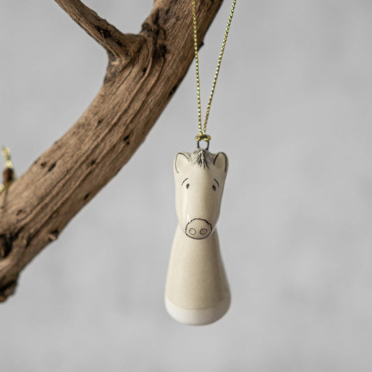 A white ceramic horse ornament with black painted eyes and a small mouth. The decoration is white with black markings and is hanging from a gold string.