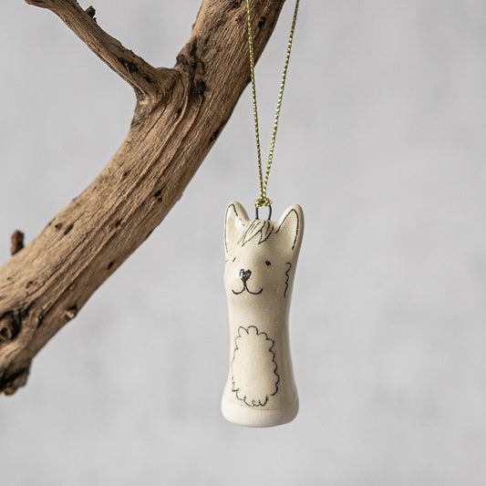 A white ceramic llama ornament with black painted eyes and a smiling mouth. The decoration is white with black markings and is hanging from a gold string.
