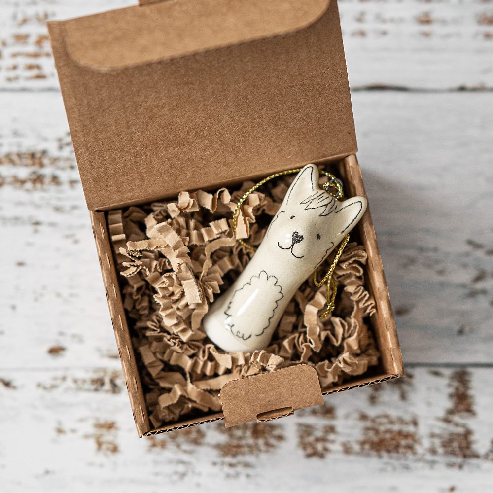 A white ceramic llama ornament with black painted eyes and a smiling mouth. The decoration is white with black markings and is hanging from a gold string.