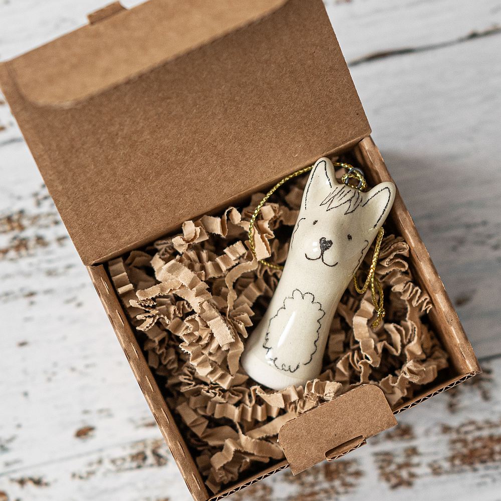 A white ceramic llama ornament with black painted eyes and a smiling mouth. The decoration is white with black markings and is hanging from a gold string.