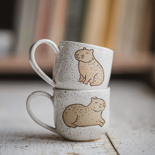 Handmade white ceramic mugs with a matte glaze, featuring an unglazed sitting capybara design on the side of one cup and lying capybara on another.