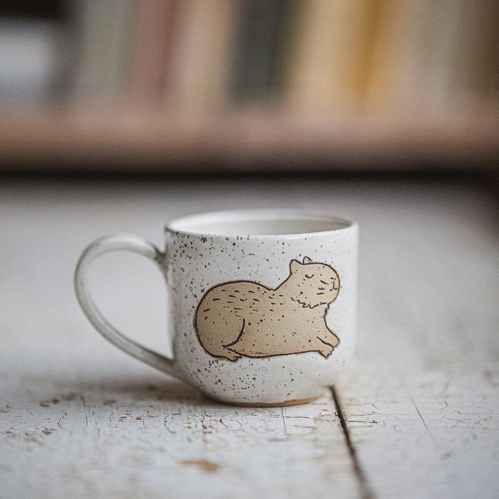 Handmade white ceramic mug with a matte glaze, featuring an unglazed lying capybara design on the side.