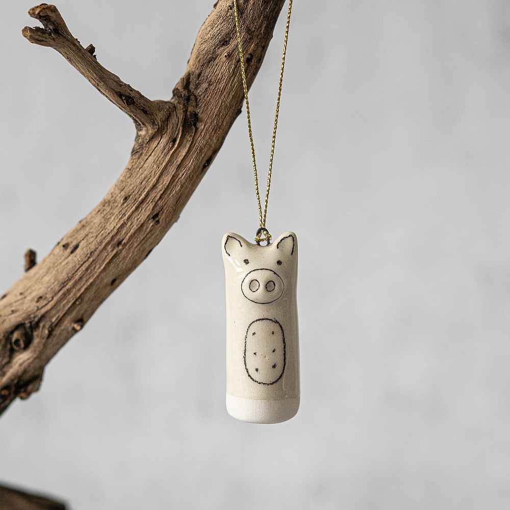 A white ceramic pig ornament with black painted eyes and a nose. The decoration is white with black markings and is hanging from a gold string.
