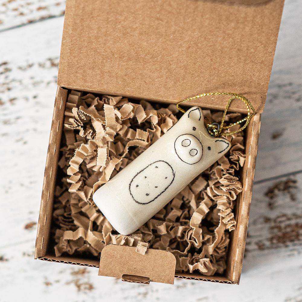 A white ceramic pig ornament with black painted eyes and a nose. The decoration is white with black markings and is hanging from a gold string.