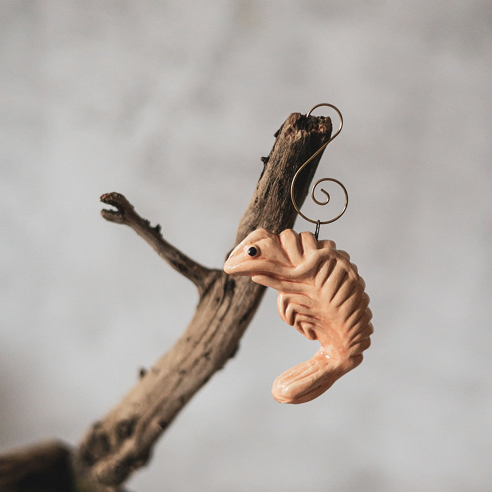 A ceramic pink shrimp ornament with black eyes. The decoration is handmade and comes with yellow hook.
