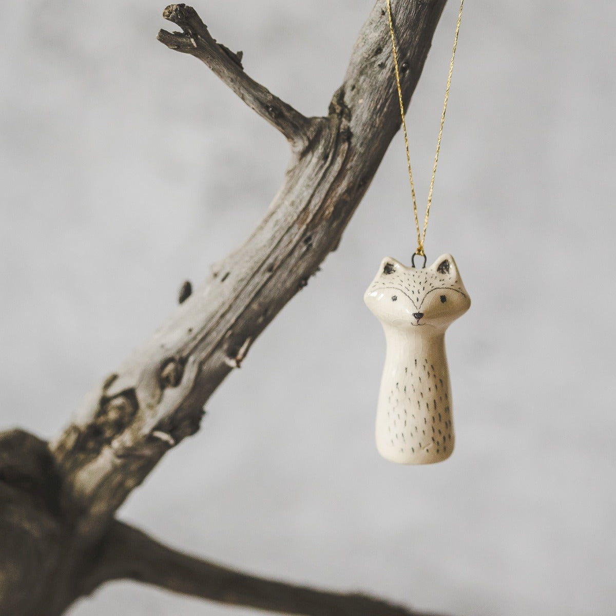 A ceramic wild raccoon ornament with hand-drawn black eyes and a black nose. The decoration is white with black markings and is hanging from a gold string.