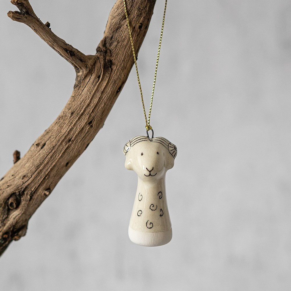 A white ceramic sheep ornament with black painted eyes and a small mouth. The decoration is white with black markings and is hanging from a gold string.