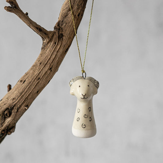 A white ceramic sheep ornament with black painted eyes and a small mouth. The decoration is white with black markings and is hanging from a gold string.