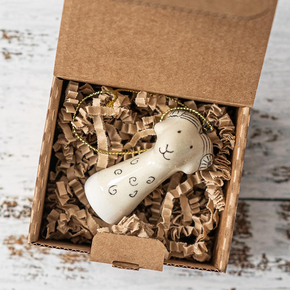 A white ceramic sheep ornament with black painted eyes and a small mouth. The decoration is white with black markings and is hanging from a gold string.