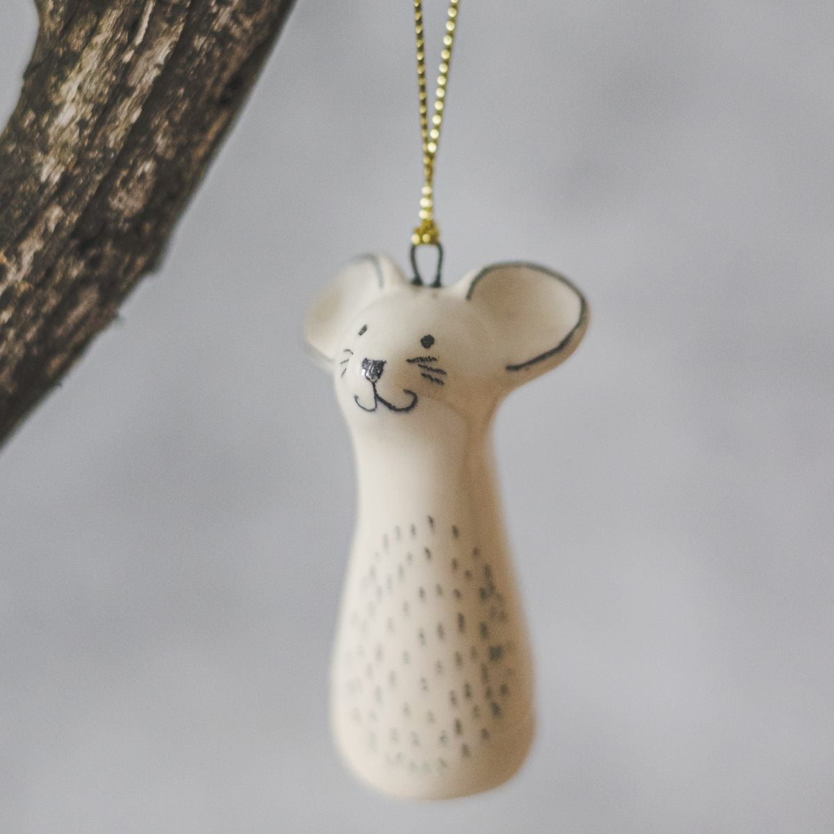A smiling ceramic mouse ornament with hand-drawn black eyes, mouth and a black nose. The decoration is white with black markings and is hanging from a gold string.