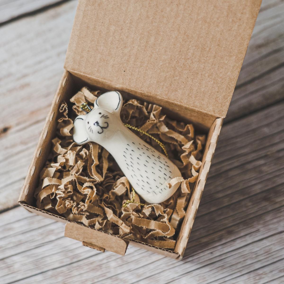 A smiling ceramic mouse ornament with hand-drawn black eyes, mouth and a black nose. The decoration is white with black markings and is hanging from a gold string.