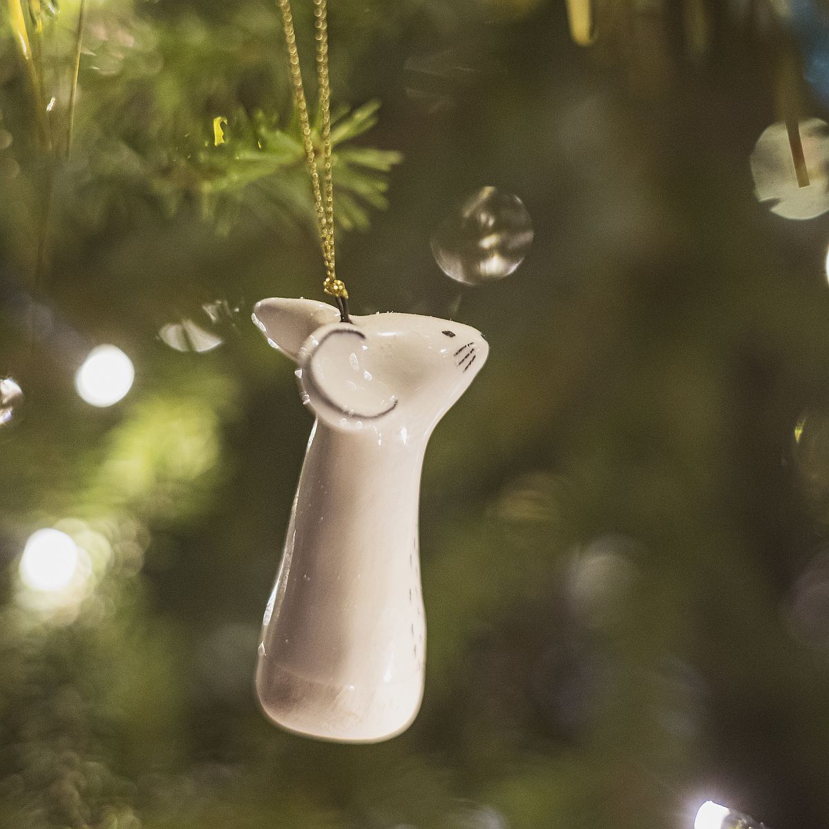 A smiling ceramic mouse ornament with hand-drawn black eyes, mouth and a black nose. The decoration is white with black markings and is hanging from a gold string.