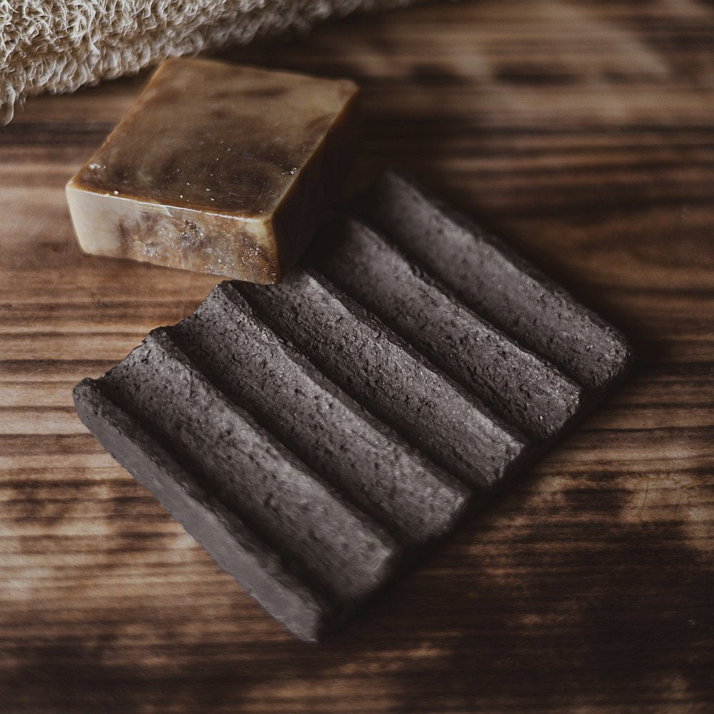 The soap dish is made of black grog clay, which gives it a textured, earthy appearance. The surface features evenly spaced grooves that run across the dish, serving as drainage channels to keep the soap dry and extend its life.