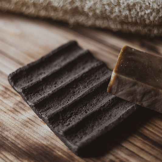 The soap dish is made of black grog clay, which gives it a textured, earthy appearance. The surface features evenly spaced grooves that run across the dish, serving as drainage channels to keep the soap dry and extend its life.