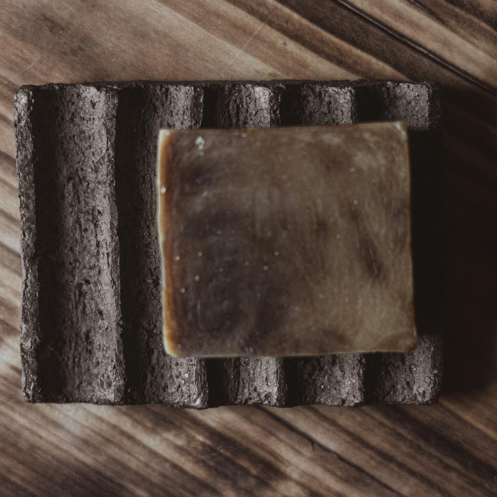 The soap dish is made of black grog clay, which gives it a textured, earthy appearance. The surface features evenly spaced grooves that run across the dish, serving as drainage channels to keep the soap dry and extend its life.