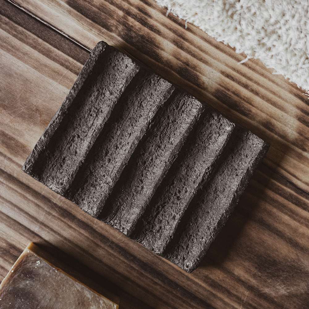 The soap dish is made of black grog clay, which gives it a textured, earthy appearance. The surface features evenly spaced grooves that run across the dish, serving as drainage channels to keep the soap dry and extend its life.