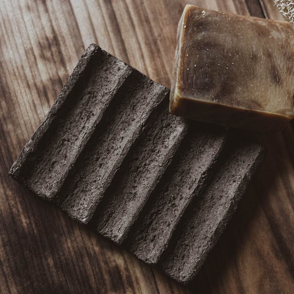 The soap dish is made of black grog clay, which gives it a textured, earthy appearance. The surface features evenly spaced grooves that run across the dish, serving as drainage channels to keep the soap dry and extend its life.