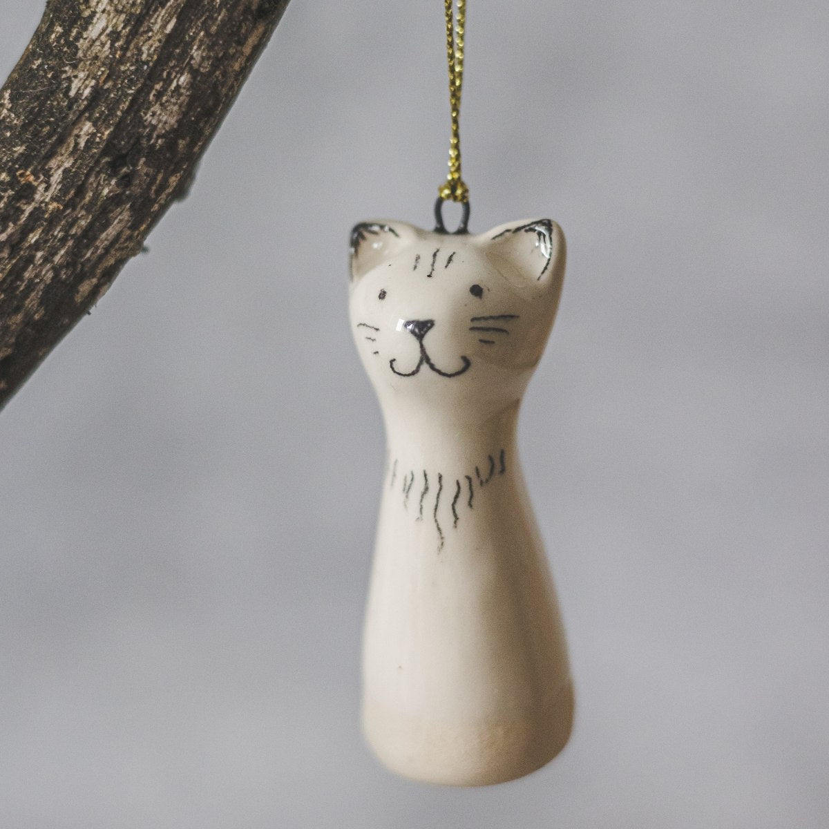 A smiling ceramic cat ornament with hand-drawn black eyes, mouth and a black nose. The decoration is white with black markings and is hanging from a gold string.