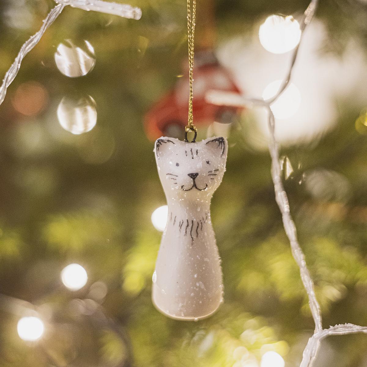 A smiling ceramic cat ornament with hand-drawn black eyes, mouth and a black nose. The decoration is white with black markings and is hanging from a gold string.