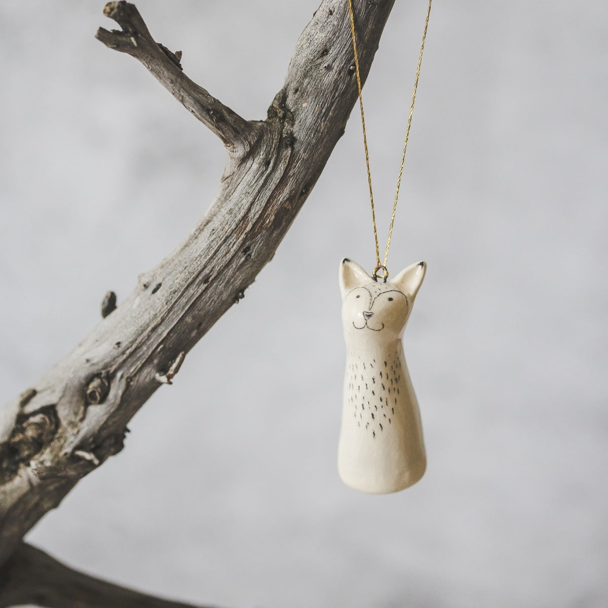 A smiling ceramic fox ornament with hand-drawn black eyes, mouth and a belly. The decoration is white with black markings and is hanging from a gold string.