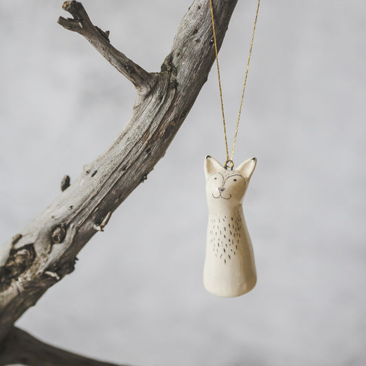 A smiling ceramic fox ornament with hand-drawn black eyes, mouth and a belly. The decoration is white with black markings and is hanging from a gold string.