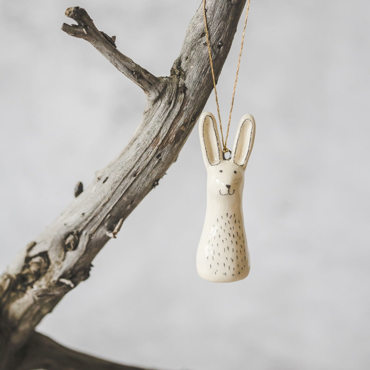 A smiling ceramic hare ornament with hand-drawn black eyes, belly and a black nose. The decoration is white with black markings and is hanging from a gold string.