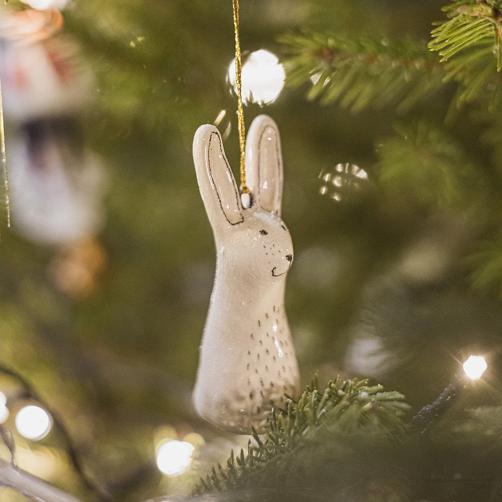 A smiling ceramic hare ornament with hand-drawn black eyes, belly and a black nose. The decoration is white with black markings and is hanging from a gold string.